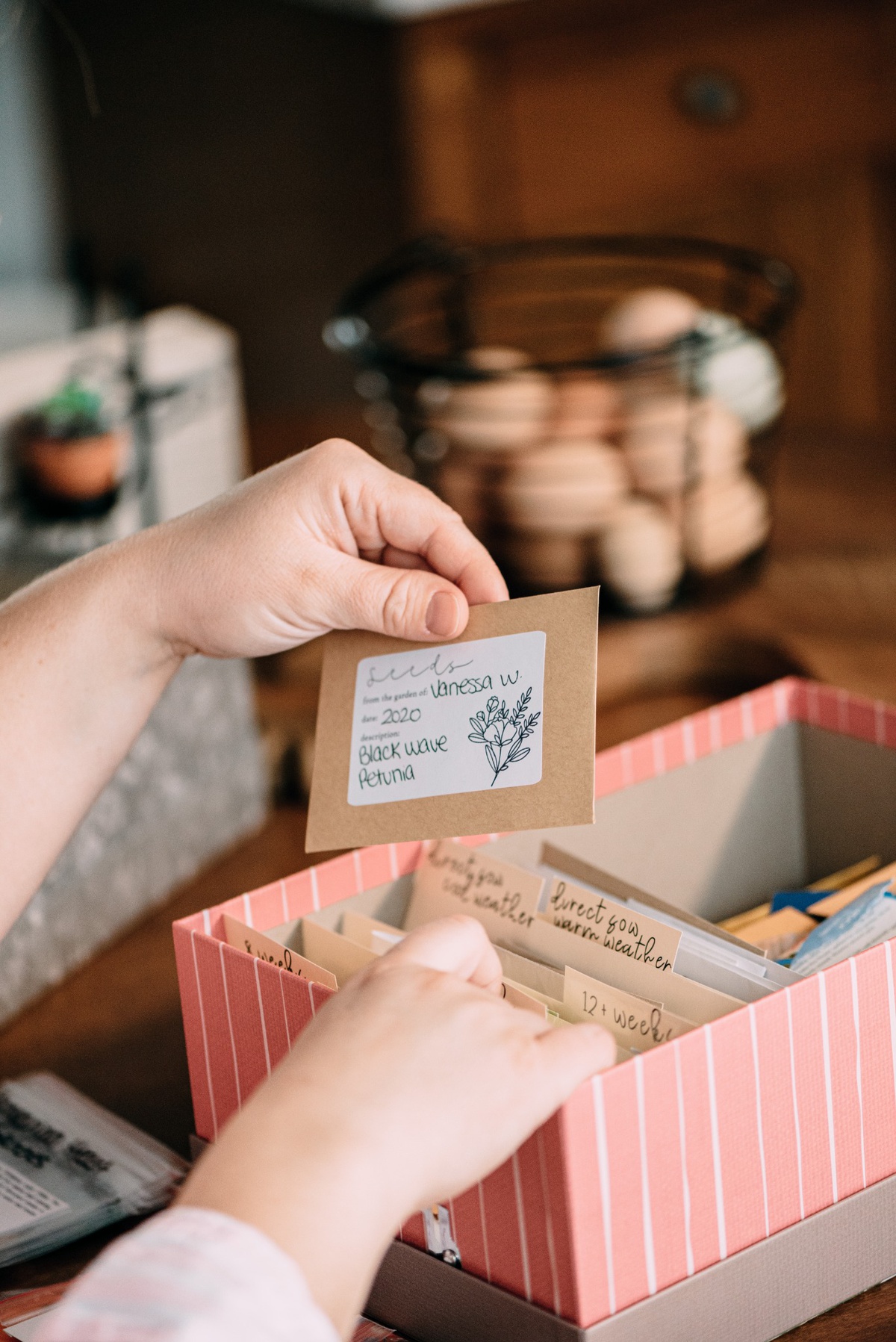 A hand holding a personalized seed packet with handwritten details, above a pink-striped box with more seed packets organized within.