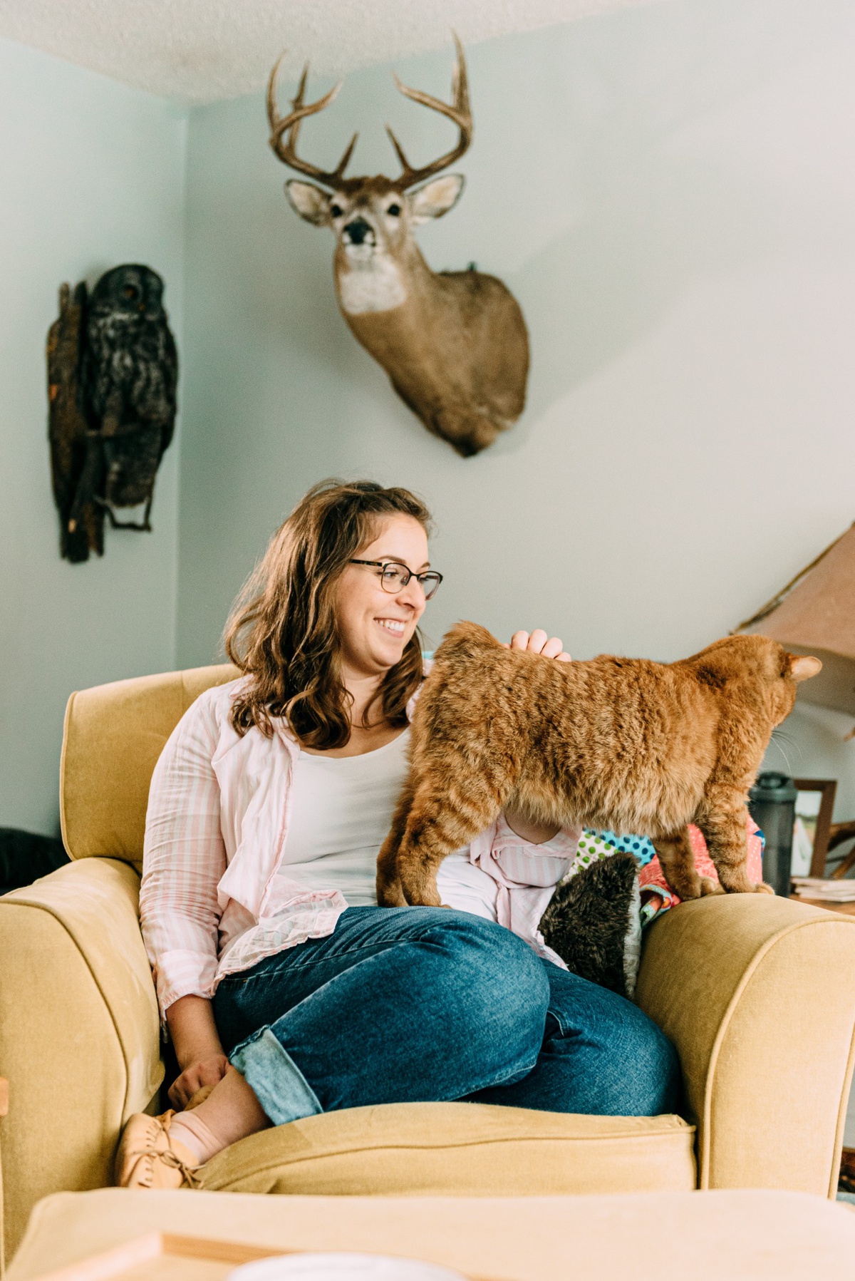 A smiling woman in a cozy indoor setting, interacting with a cat, with rustic decor that includes a mounted deer head on the wall.