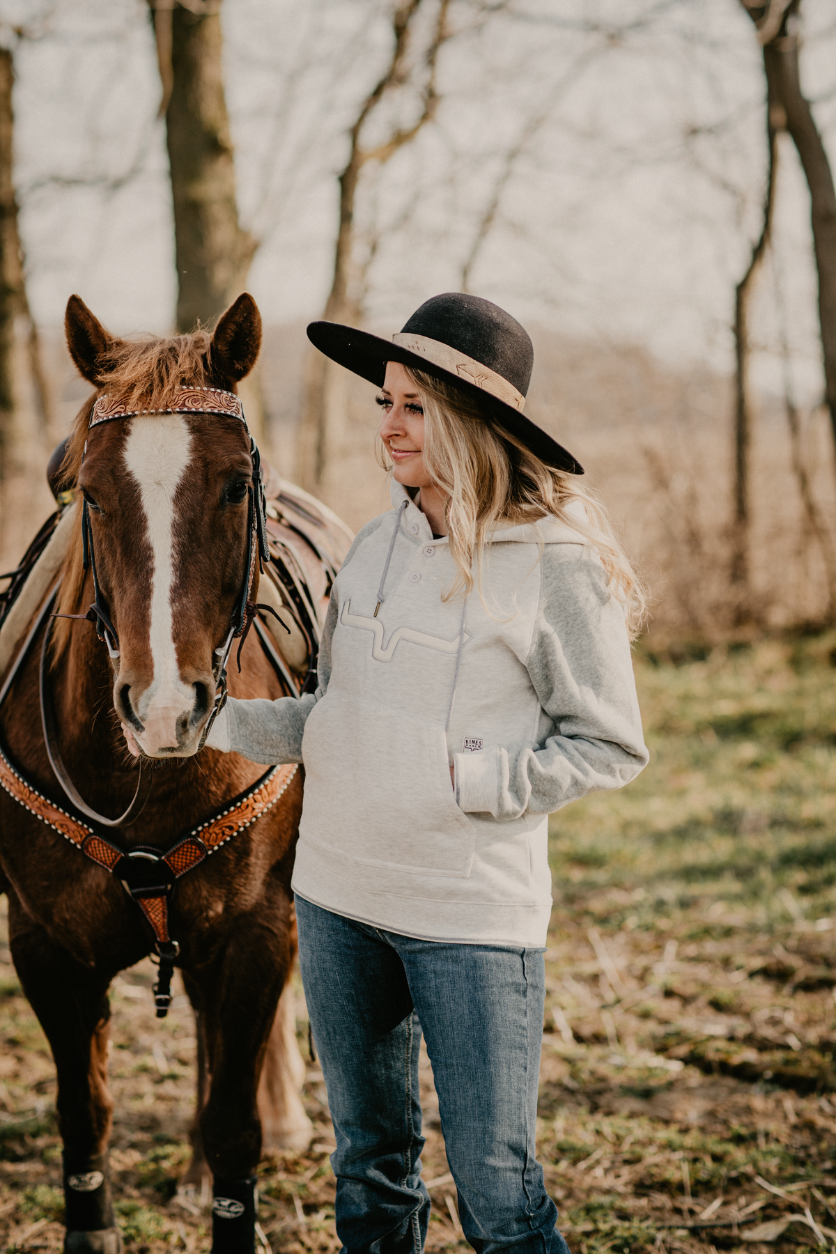 A smiling woman in a black hat stands next to a bay horse, dressed in a casual grey hoodie, embodying a relaxed equestrian style.