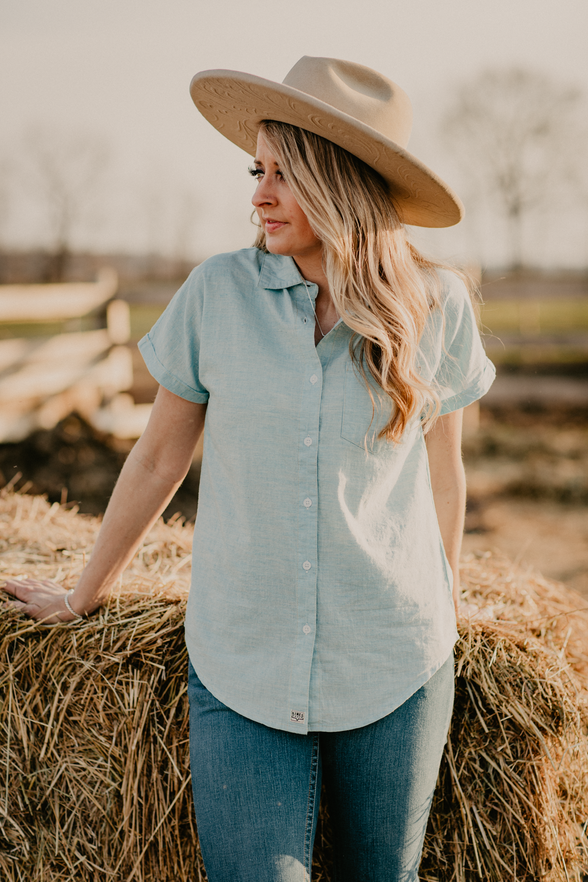 A stylish woman leans on a hay bale, wearing a light blue shirt and jeans, paired with a tan cowboy hat, exuding a chic farm-inspired look.