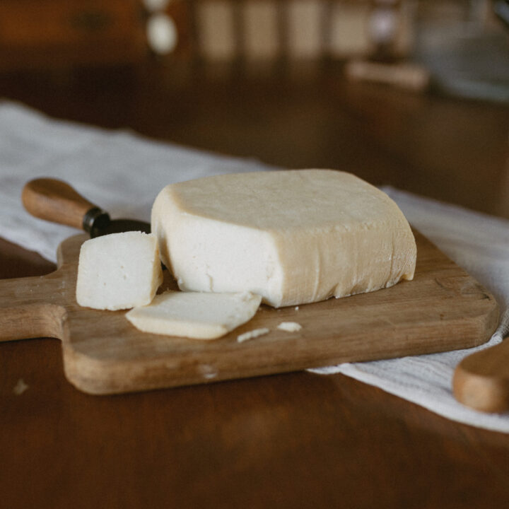 A large wheel of homemade cheese on a wooden cutting board, with a cheese knife and a few slices cut off to the side.