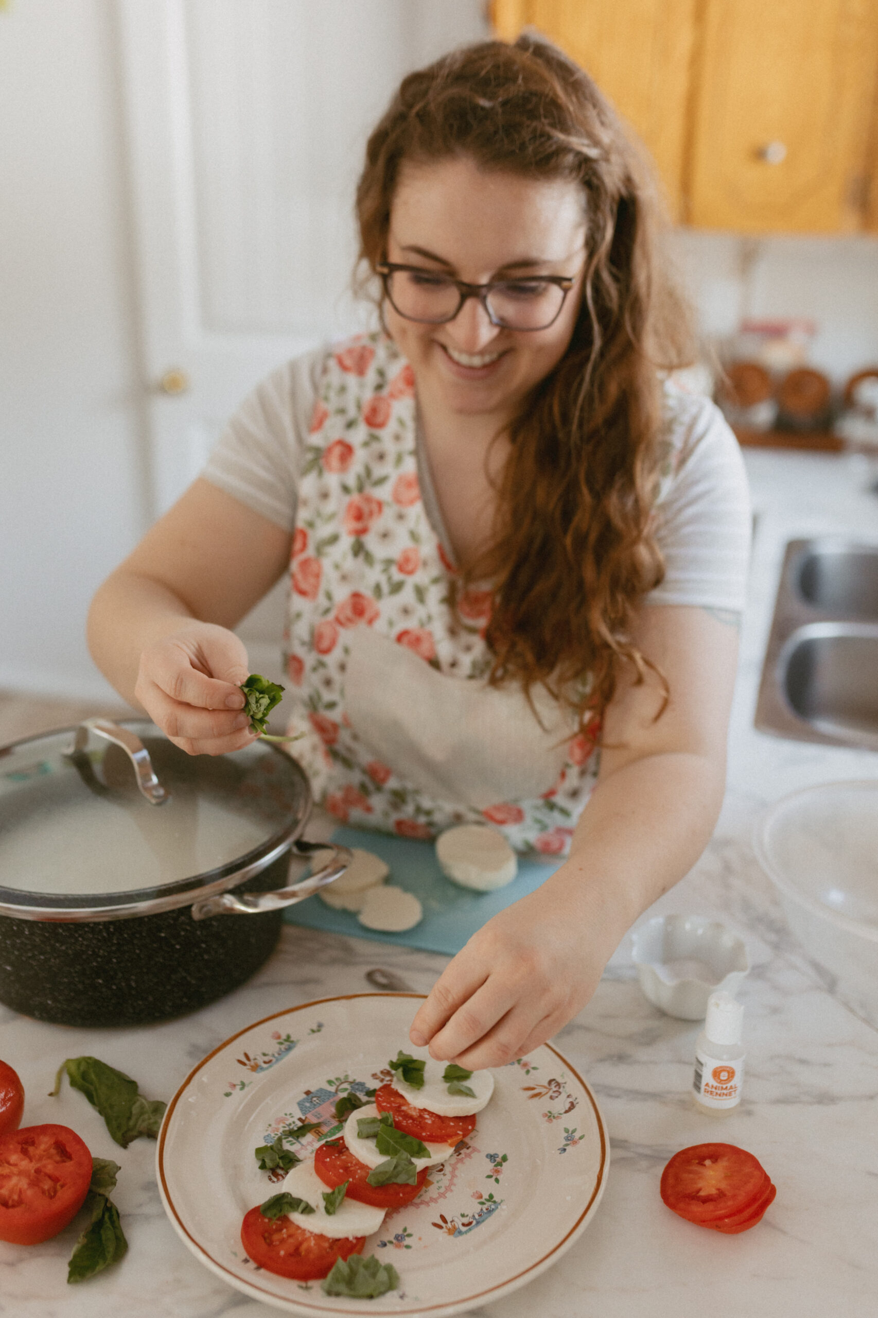 A woman assembles a caprese salad with freshly made cheese, basil, and tomato slices on a decorative plate, exemplifying a homemade dish with the cheese.