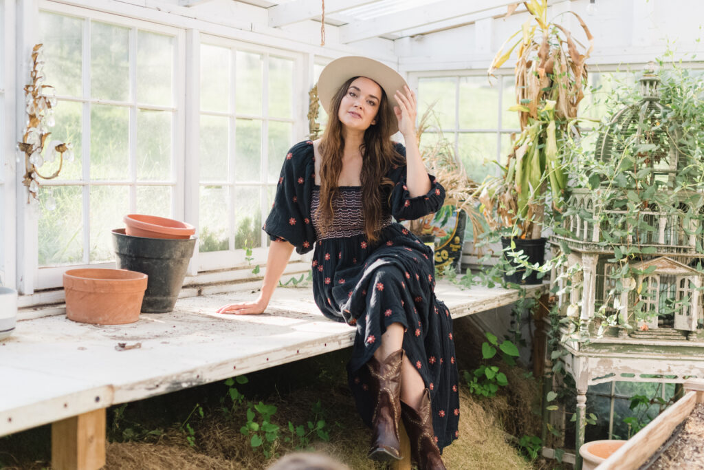woman sitting on a table in a greenhouse wearing a brimmed hat and a black dress with cowboy boots