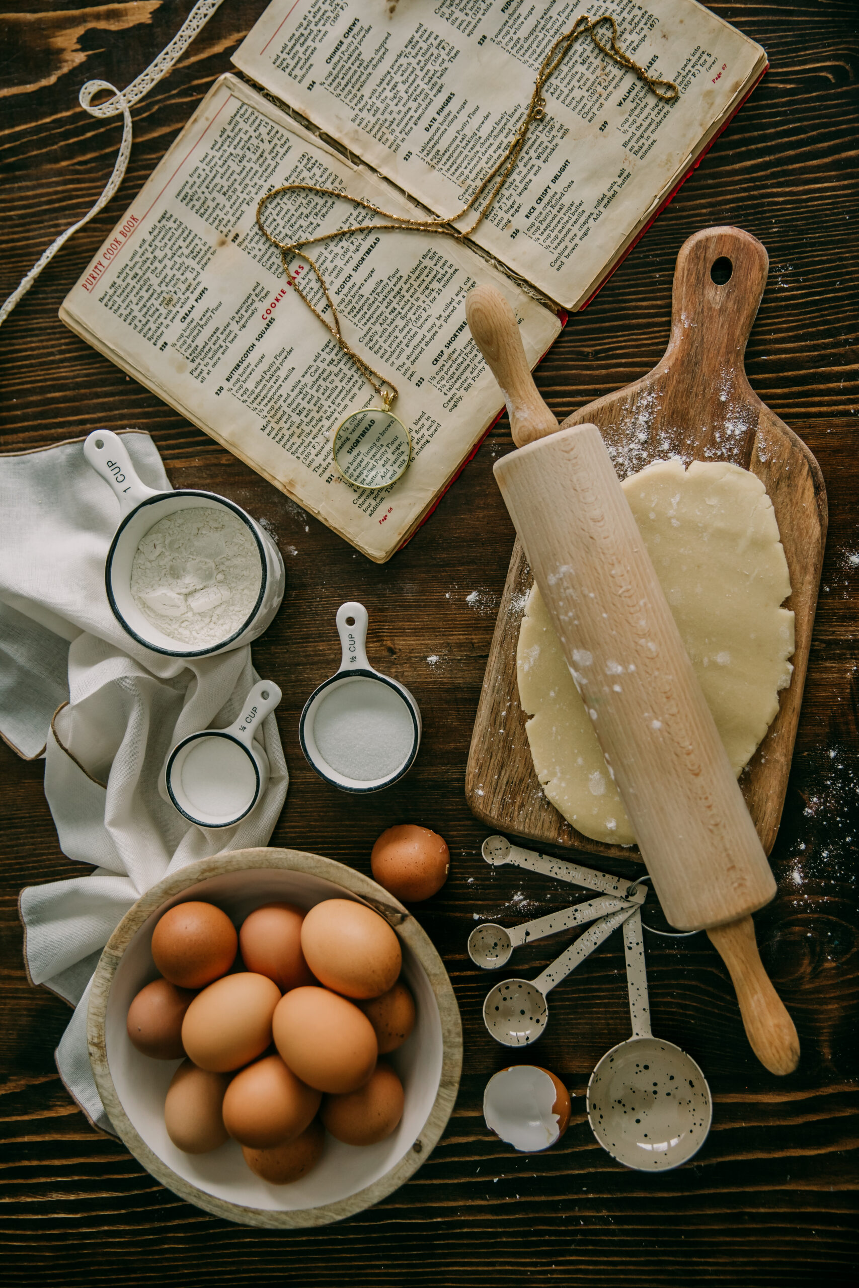 Top view of a wooden table with baking ingredients and tools including a rolling pin, dough, eggs, measuring cups, and an open cookbook with a magnifying glass on it, set in a warm, rustic kitchen.
