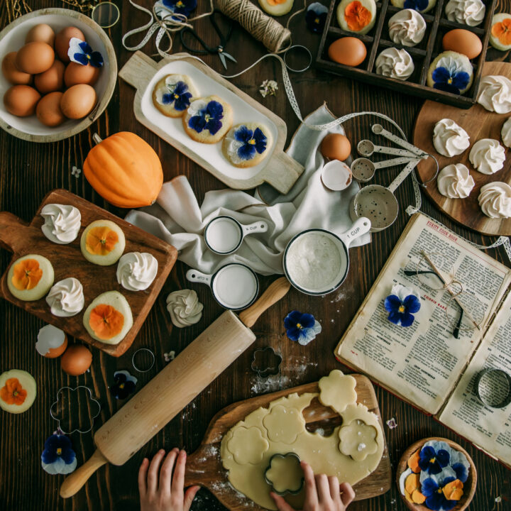 An overhead view of a rustic baking scene with hands rolling out shortbread dough on a wooden board. The table is scattered with baking ingredients and tools including eggs, a pumpkin, measuring cups, a rolling pin, and fresh pansies. Some of the finished products, vanilla meringues and pansy-topped shortbread cookies, are displayed on trays. An open cookbook adds a vintage touch to the scene.