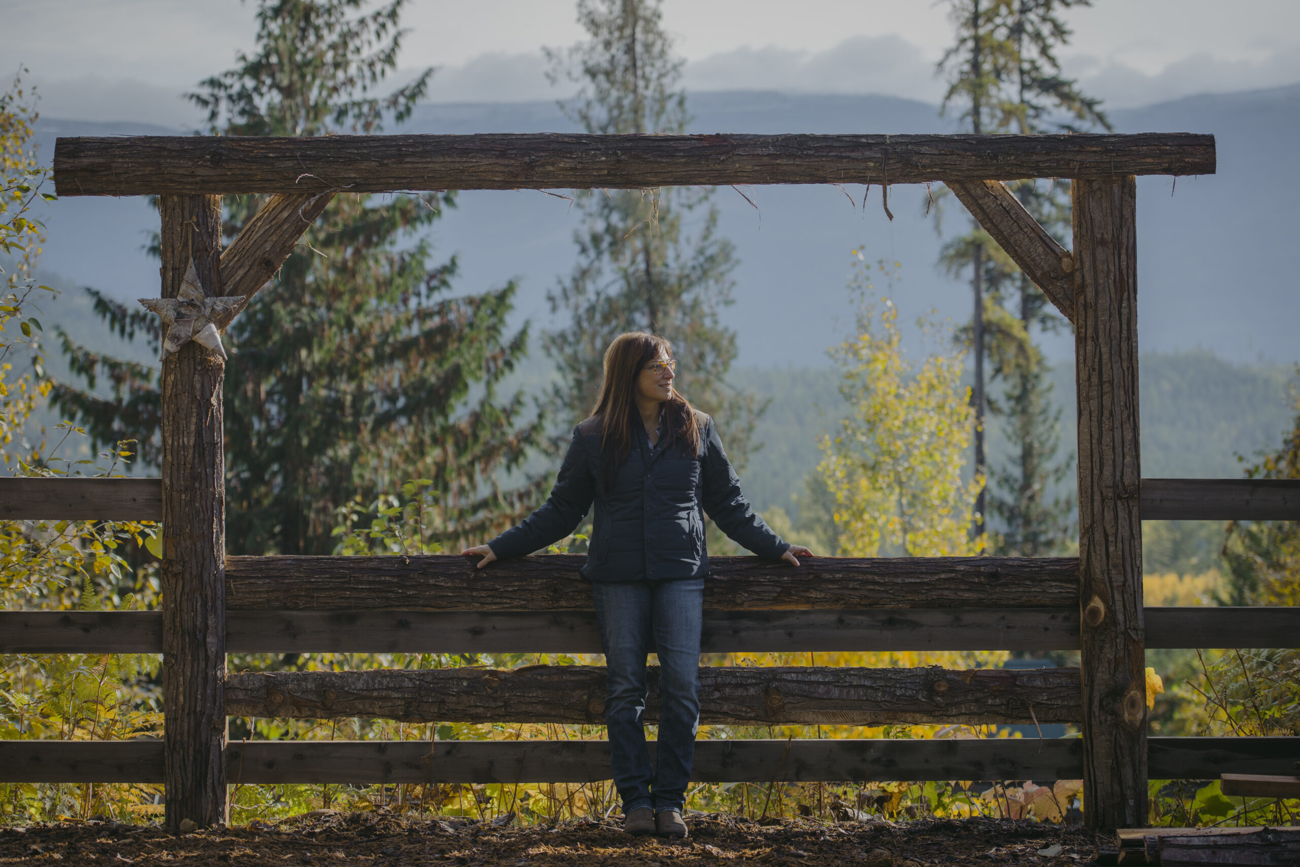 A woman leaning on a wooden fence with trees and mountains in the background.