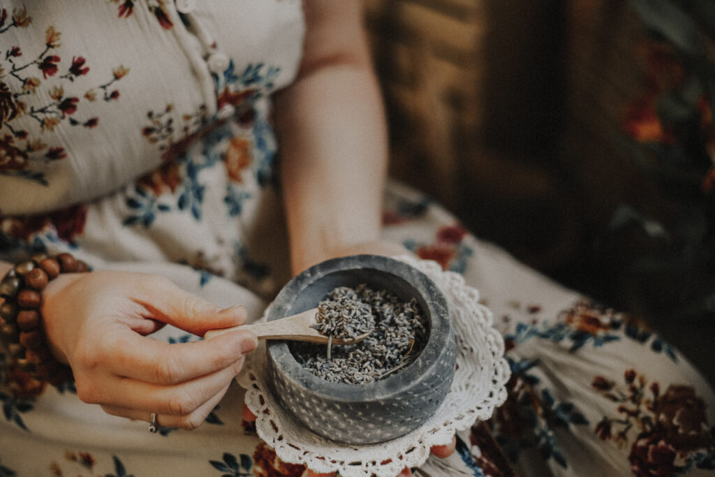 A person's hands holding a spoon and a mortar and pestle with lavender buds on a lace cloth.