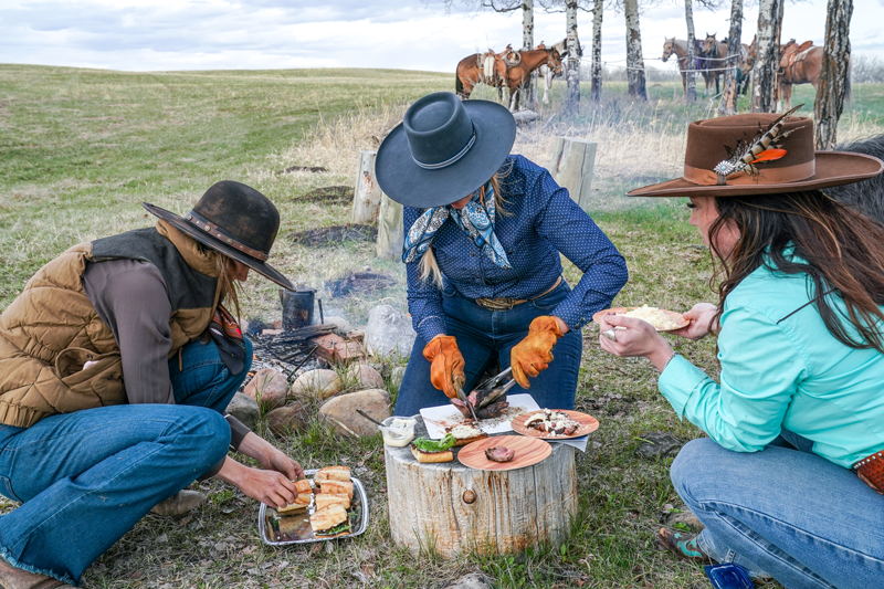 3 women in cowboy hats crouch down around a tree stump. The middle woman is cutting steak