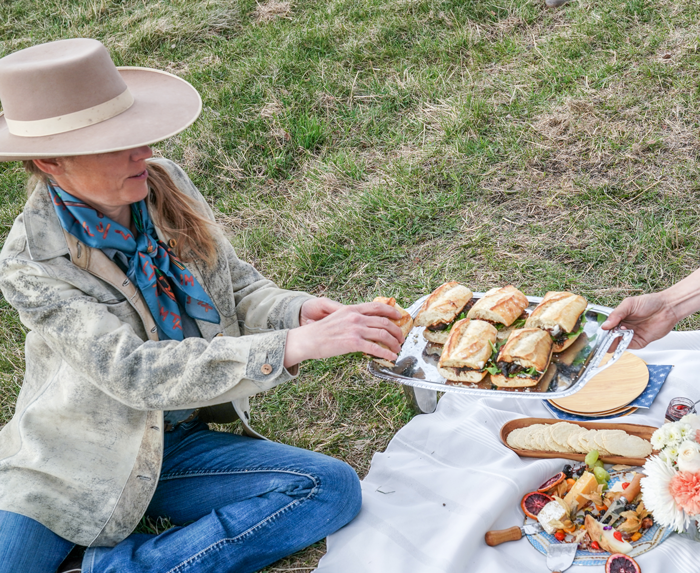 Woman in cowboy hat takes a sandwich off a platter above a white blanket and other food on the blanket.