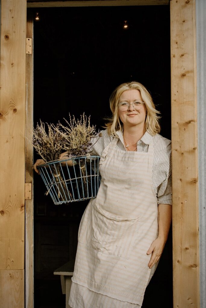 A woman in a light blue shirt and a cream coloured apron holding a basket of lavender leaning against a door frame