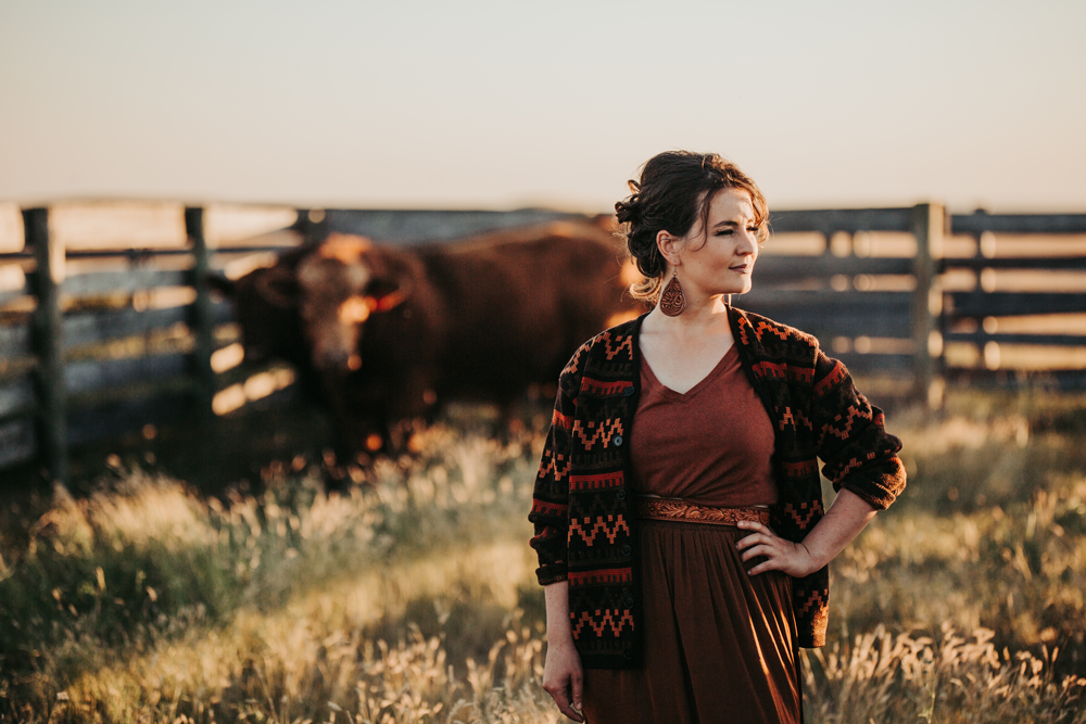 woman stands in a field of grass with a fence and cow behind her