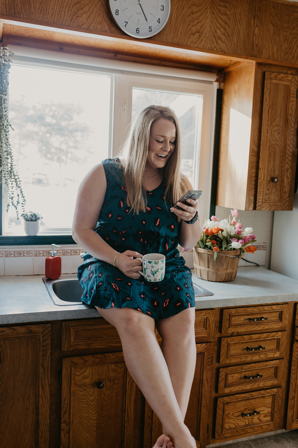 Woman in teal dress sits on counter in kitchen with a coffee mug in one hand and a phone in the other, looking at the phone