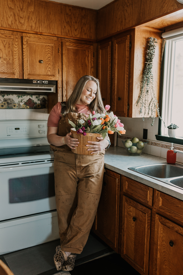 Woman in brown overalls leans against the counter in a kitchen with a basket of tulips in her hands.