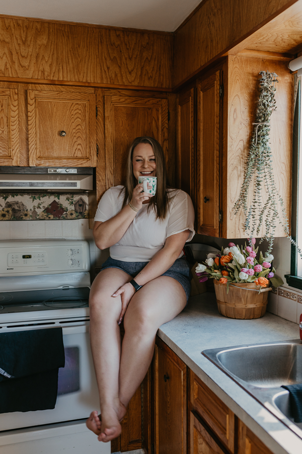 Woman in white tshirt and blue denim shorts sits on the counter in a kitchen with a coffee mug and a big smile