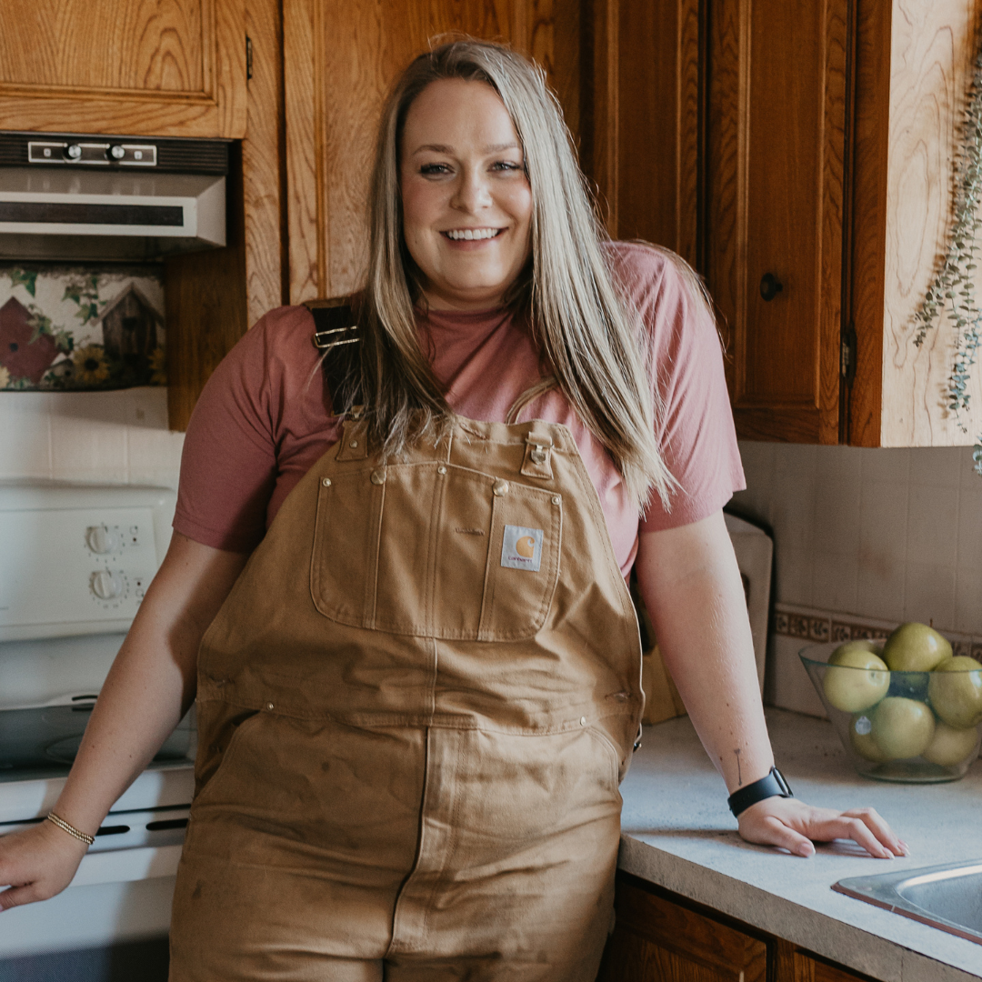 Woman in brown overalls leans against the counter in a kitchen