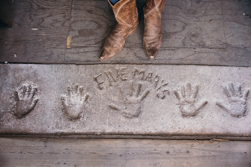 a pair of brown cowboy boots stand above a concrete slab with hand prints and five marys written in it