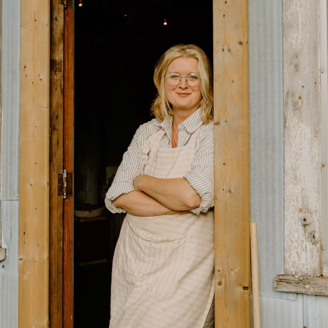 Woman in light blue shirt and cream apron stands with arms crossed leaning against a door frame