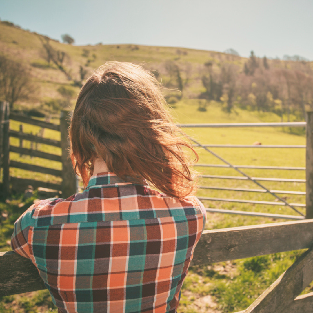 Back of a woman in a plaid shirt looking out into a field holding onto a rail from a fence