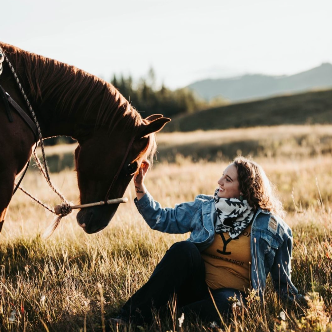 woman sits in grass rubbing the head of a brown horse