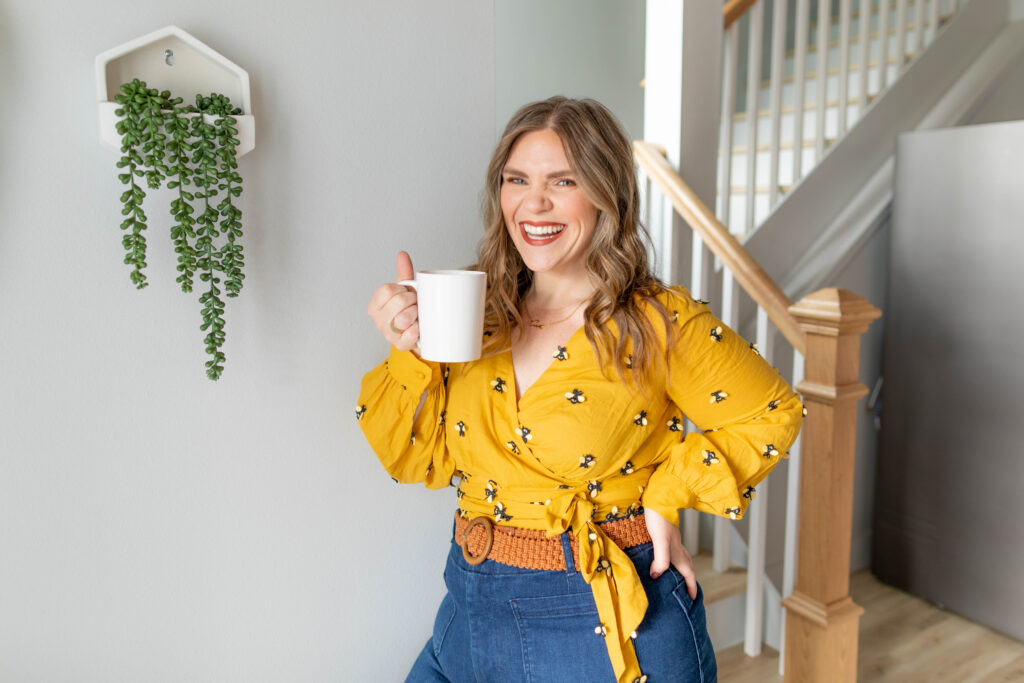 Woman in mustard coloured blouse and jeans with a white coffee mug in her hand standing in front of a staircase