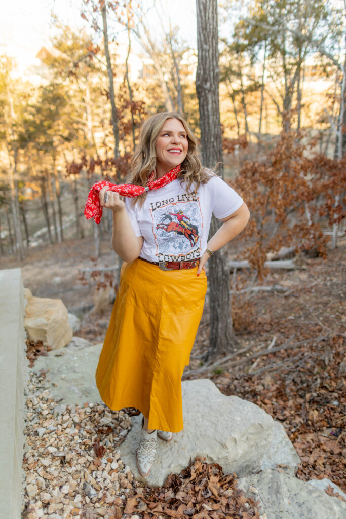 Woman in a white tee, mustard ankle length skirt and red scarf standing on a rock in the forest.