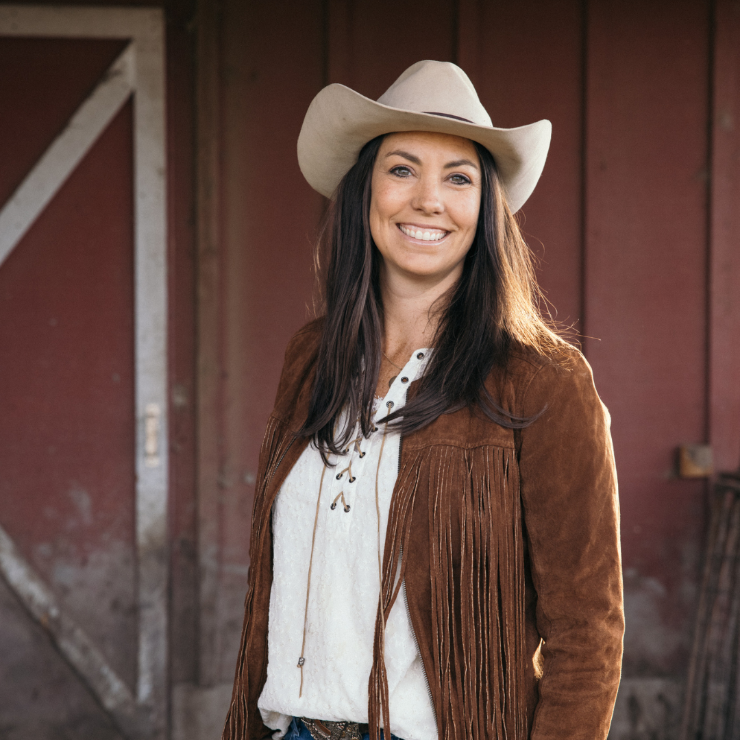 woman in brown jacket and white cowboy hat, smiles and stands in front of red barn
