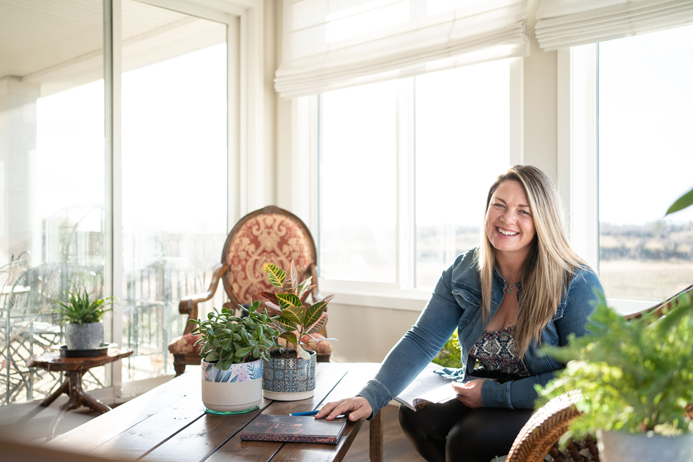 Woman sits at table with her hand on the table with plants around and lots of windows behind her