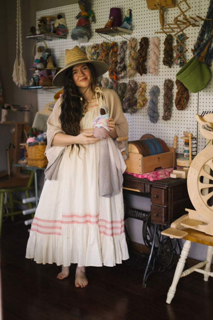 Woman in a white dress and hat standing in a room with dark floors and a white peg board behind her filled with wool