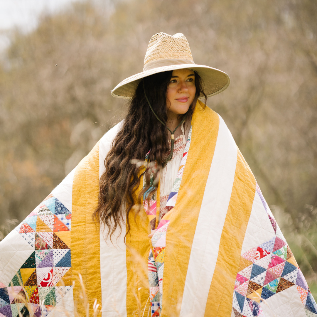 woman draped in a quilt with yellow and white, wearing a hat standing outside.