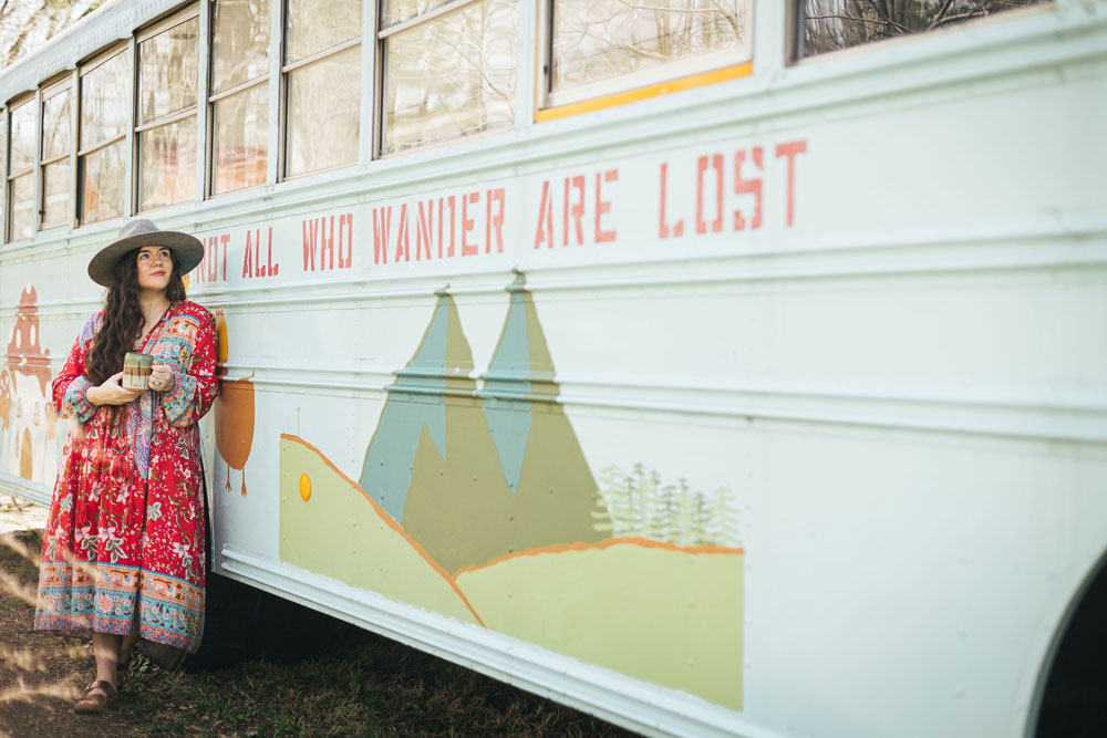 woman in red dress and hat standing beside a bus that says not all who wander are lost