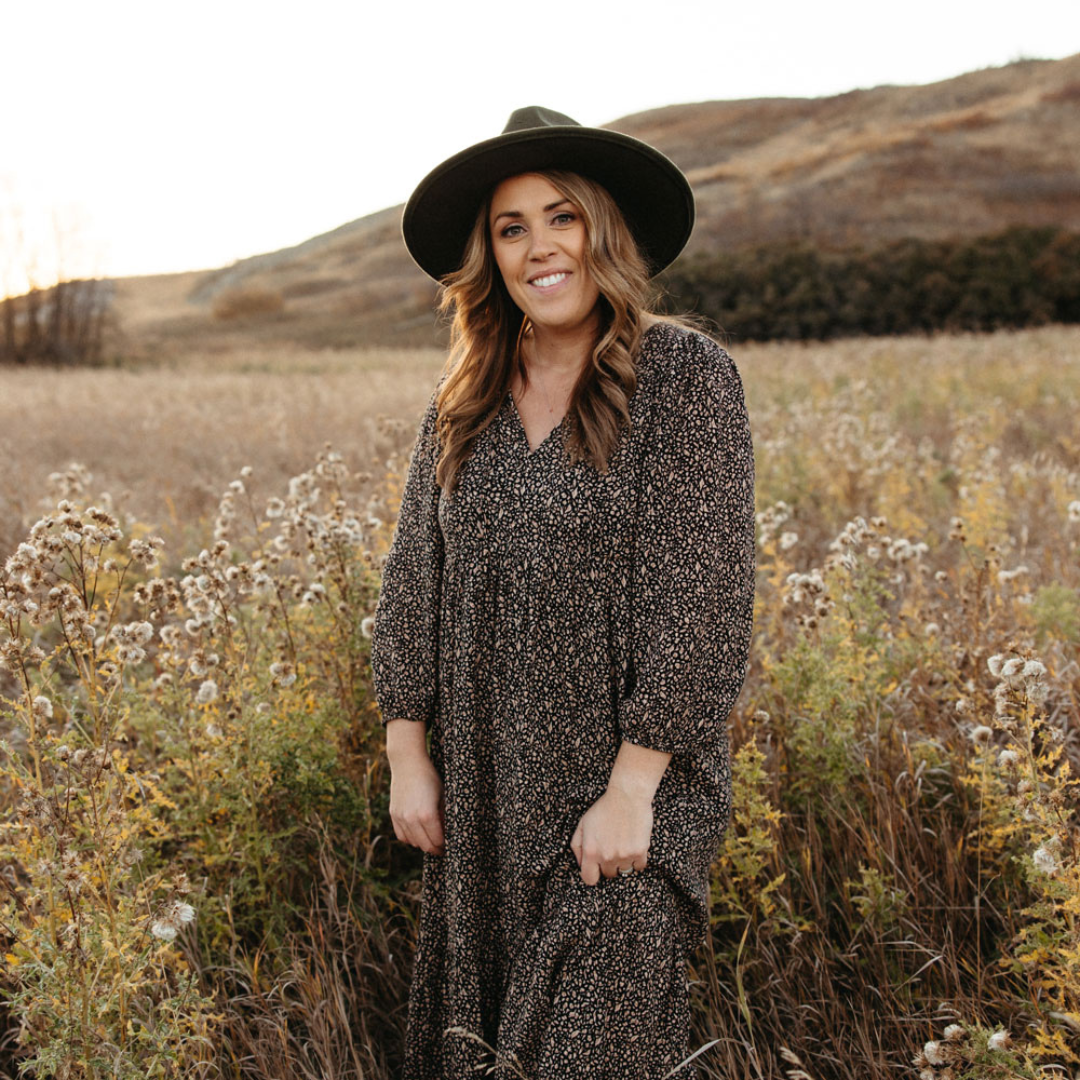 woman in brown dress and black hat stands in a field of flowers