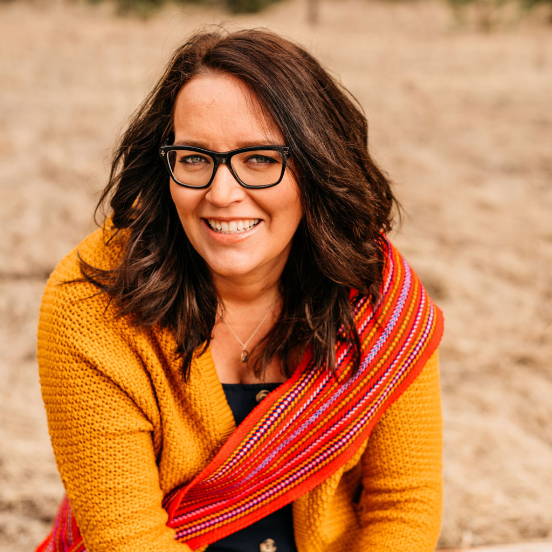 woman with dark hair and glasses sits outside wearing a golden sweater and a red indigenous scarf