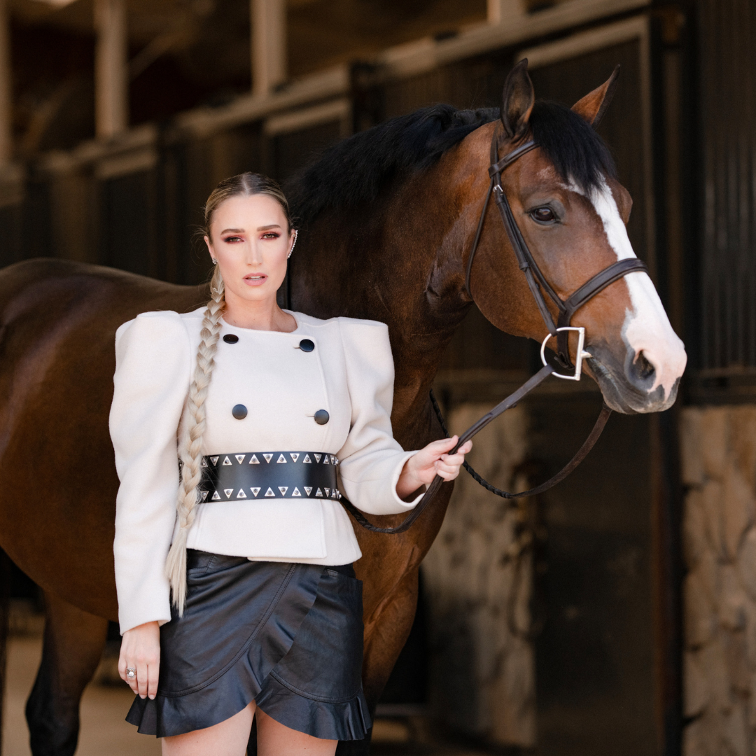 woman with a white blazer and black shorts stands beside a large brown horse