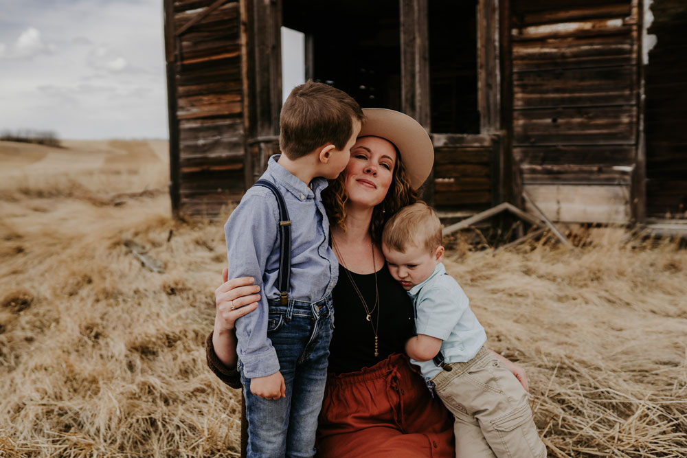 A woman hugs two young boys in front of a barn