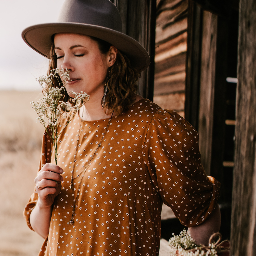 A woman in a brown hat and a dark mustard dress smells a flower