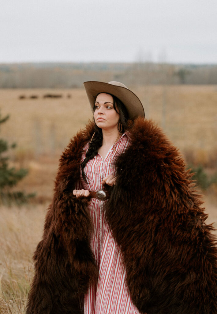 Woman in pink dress and brown hat wears a buffalo fur around her, standing outside in a field