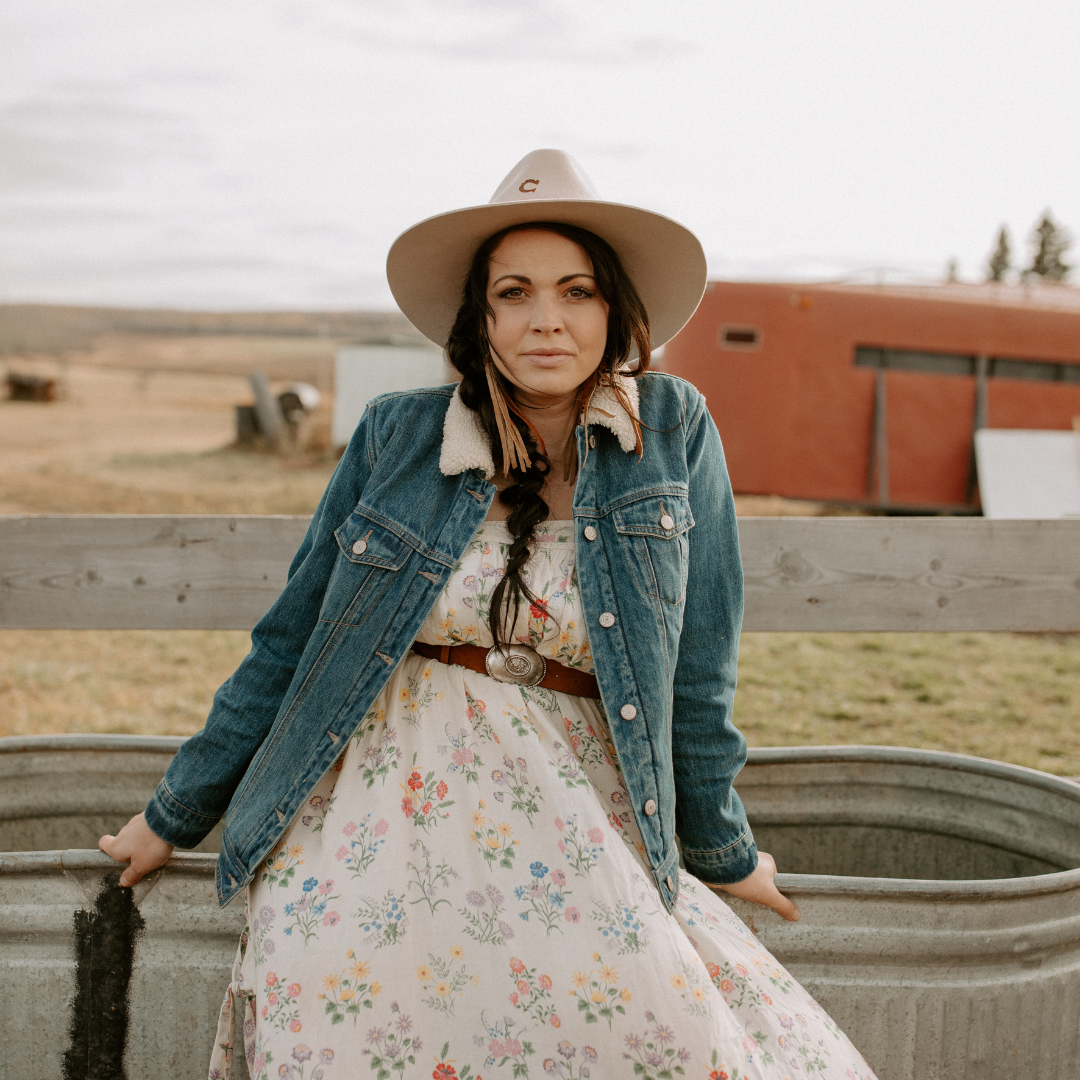 Woman in dress with a jean jacket and hat sits on the side of a fence with a barn and field in the background
