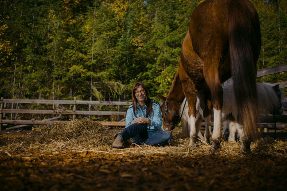 Woman sits on ground wearing blue jeans and a blue shirt beside her brown horse