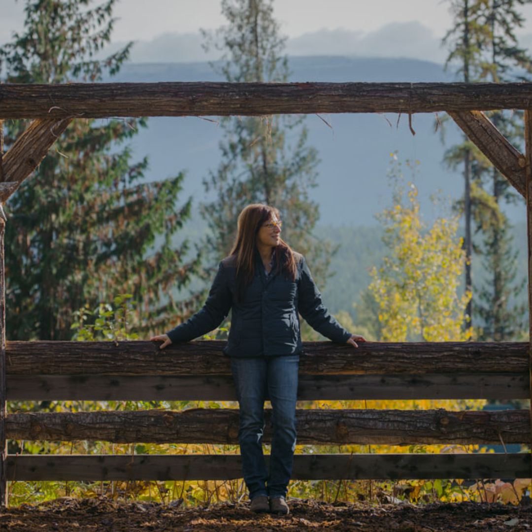 Woman stands leaning on fence with the trees and mountains in the background