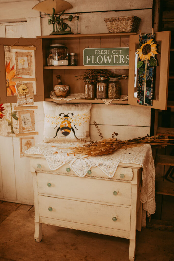 A white dresser topped with a bee pillow and some dried flowers with a shelf above