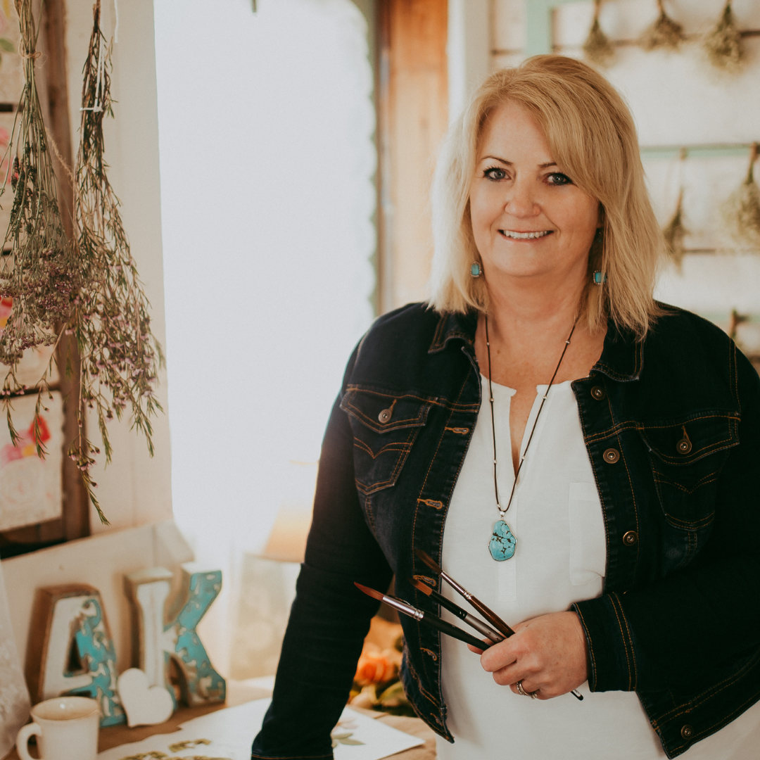 Anita stands wearing a white shirt and black blazer holding paint brushes