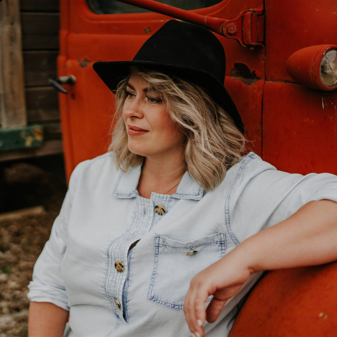 Erin sits on the ground in front of a red truck, wearing a black hat and light denim shirt