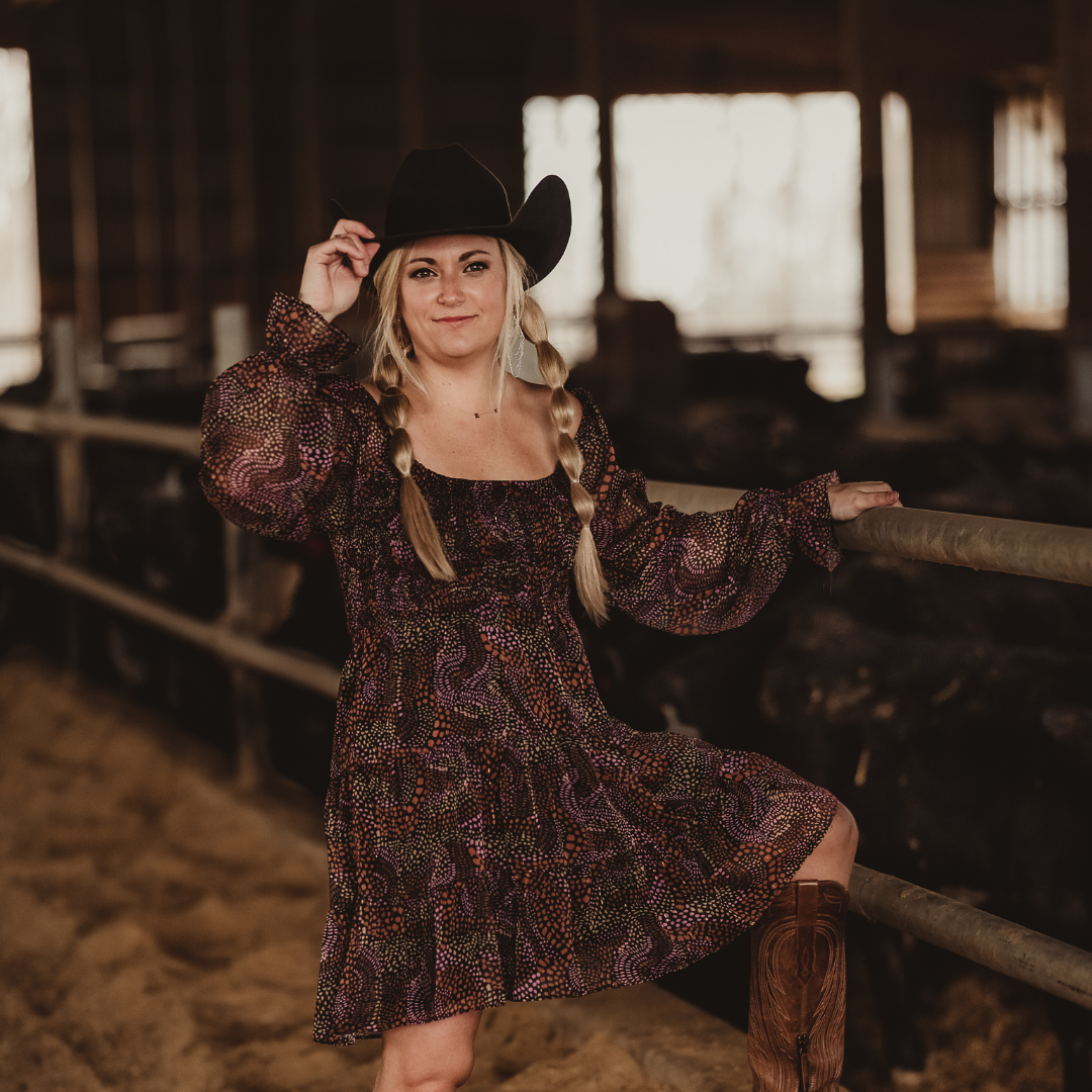 Marissa stands in her barn wearing a brown dress, brown cowboy boots and a black cowboy hat