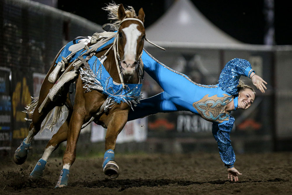 Shelby does a trick off her horse wearing a blue outfit