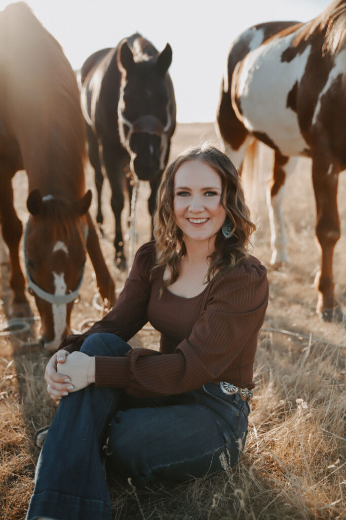 Shelby wearing a brown long sleeved top and jeans sits in a field of brown grass with 3 horses around her.