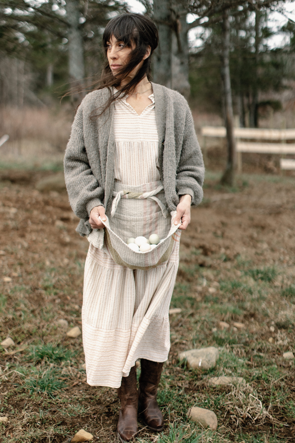 Ashley stands in a yard wearing a dress and using her apron to collect eggs