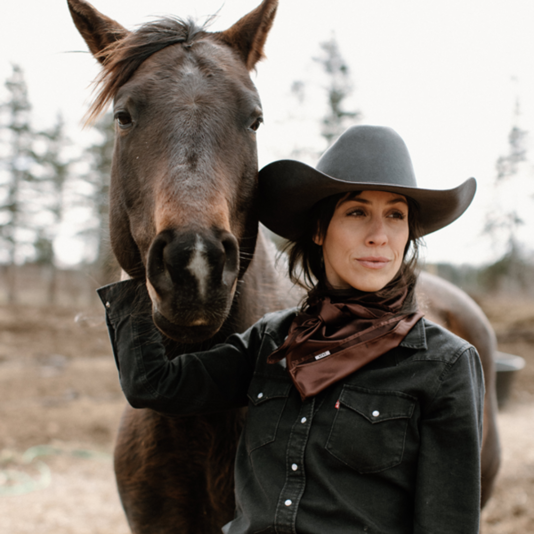 Ashley stands with her brown horse wearing a black cowboy hat, a black shirt and a brown wild rag