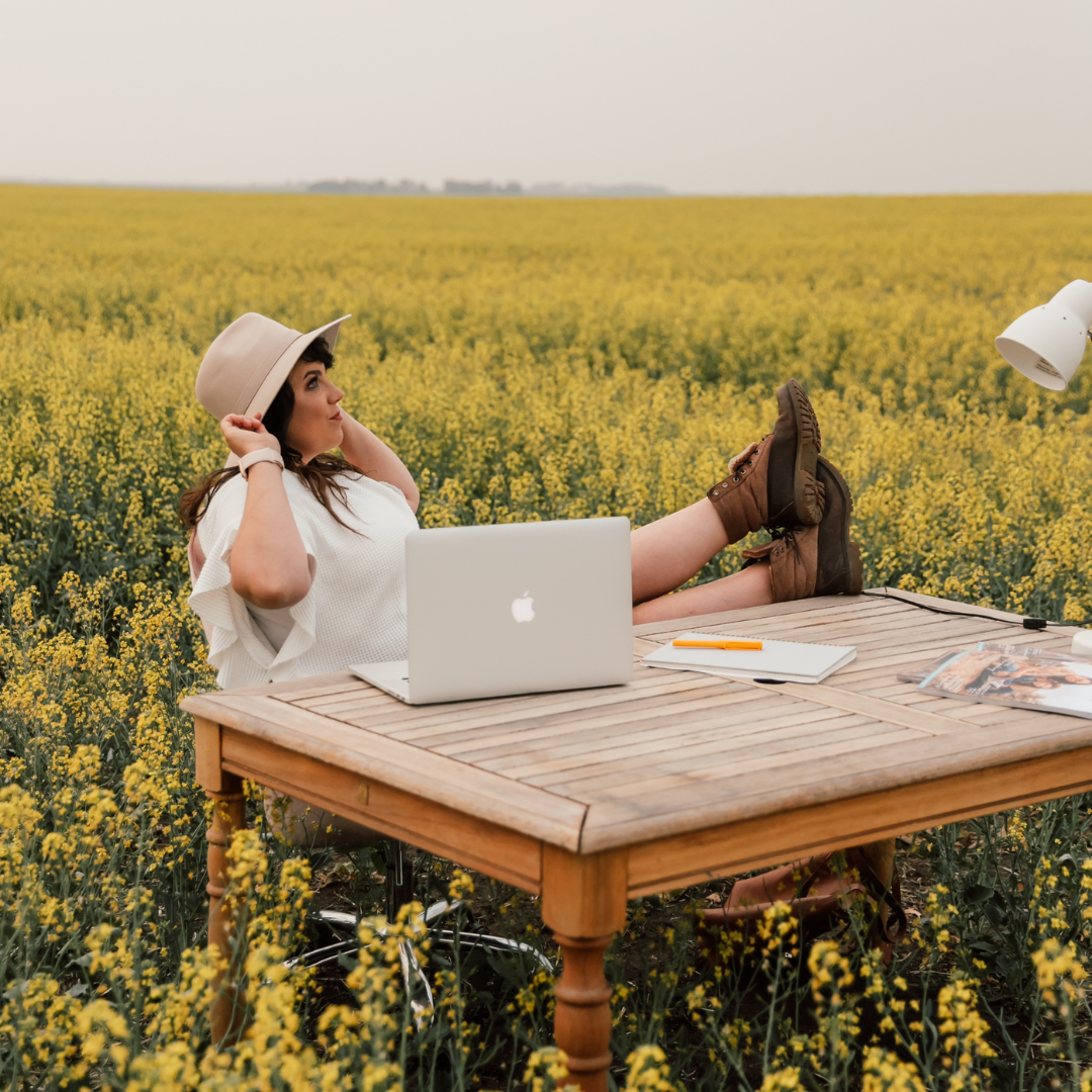 a woman sits at a desk in a field with her laptop and her feet on the desk.