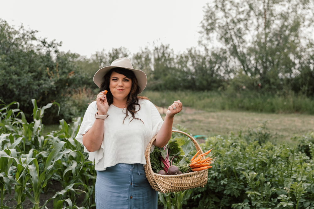 Dani stands in her garden with a basket of vegetables as she eats a carrot