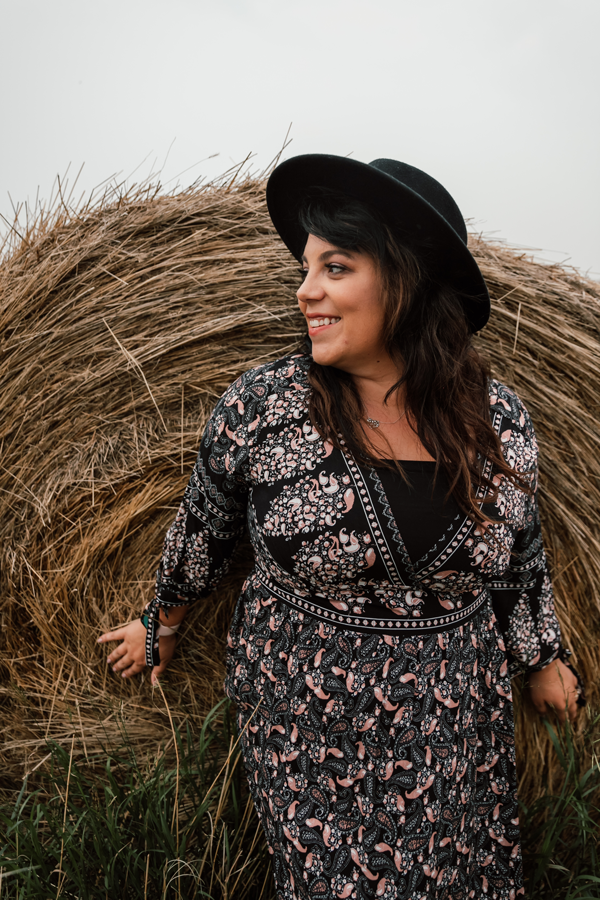 Dani wears a dress with a black hat and leans on a large hay bale